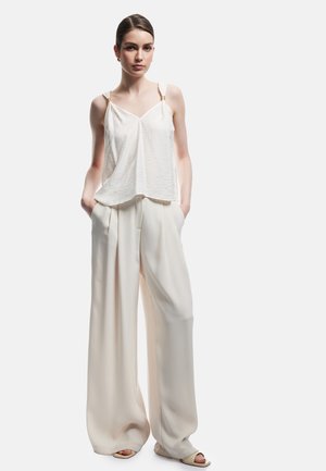 Jonge vrouw draagt een losse witte mouwloze top, brede crèmekleurige broek en sandalen met open tenen, staande met handen in de zakken.