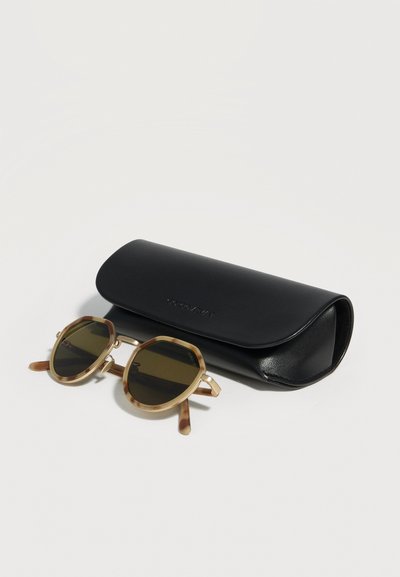 Στρογγυλά γυαλιά ηλίου με σχέδιο χελώνας και σκούρους φακούς, τοποθετημένα δίπλα σε μαύρη δερμάτινη θήκη γυαλιών ηλίου πάνω σε απλή επιφάνεια.