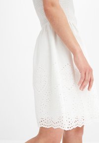 Vit bomullsklänning med en smockad bodice och en kjol som har broderade öljettmönster och en scallopad fåll. Slät textur.