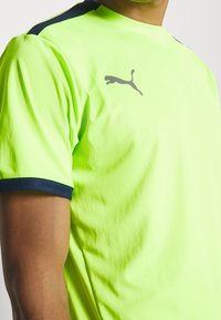 Camiseta deportiva de color amarillo neón con mangas cortas, presenta detalles en azul oscuro en los hombros y un logo gris de Puma en el pecho. Textura suave.