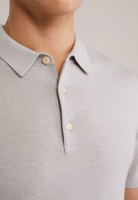 Gai pelēks adīts polo krekls ar platu apkakli, kas aprīkots ar trim bēšām pogām un gludu, teksturētu apdari. Smalkais dizains un pielāgota piegriezuma. 