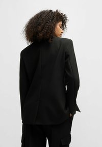 Blazer nero su misura con spalle strutturate, spacco sul retro e tessuto liscio. Abbinato a pantaloni cargo, che mostrano un design minimalista.