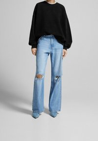 Sudadera negra de gran tamaño, jeans rasgados de color azul claro con piernas anchas, combinados con tacones de punta azul claro. Diseño y texturas minimalistas.