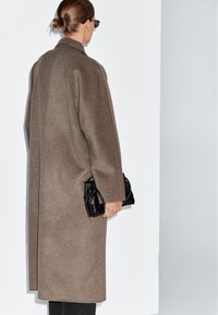 Bruine wollen jas met een ontspannen pasvorm, schuine schouders en verlaagde mouwen, gehouden met een zwarte clutch. Effen achtergrond.