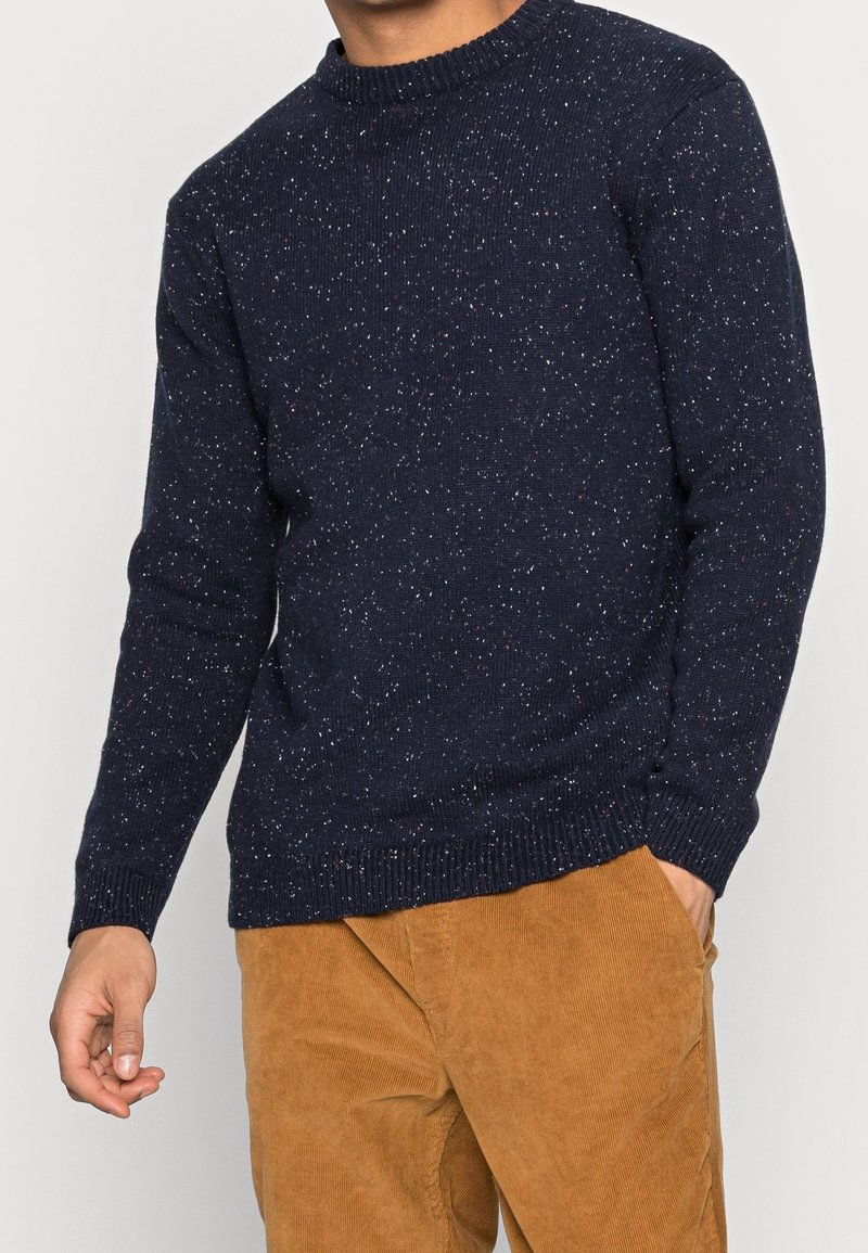 Homme portant un pull en maille bleu marine moucheté et un pantalon en velours côtelé moutarde, une main dans la poche, visible du menton aux cuisses.