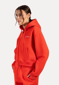 Haut hoodie rouge cropped en tissu lisse, avec une poche avant, une capuche à cordon de serrage et un logo brodé sur la poitrine gauche.