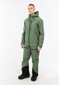 Jonge man in een groene hoodie en bijpassende groene broek met zwarte versterkte onderbenen en zwarte laarzen, staande op een witte achtergrond.