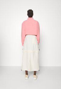 Persona con trenzas, vistiendo una chaqueta de forro polar rosa, una falda larga blanca y zapatillas beige, de pie contra un fondo blanco liso.