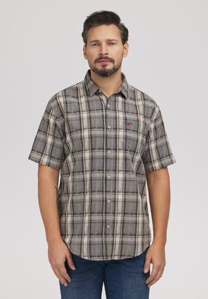 Mężczyzna o krótkich włosach i brodzie, ubrany w szarą kraciastą koszulę na guziki z krótkim rękawem oraz niebieskie dżinsy, stojący na jasnym tle.