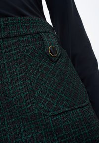 Černá látka s zeleným kostkovaným vzorem, obsahující boční kapsu s černým knoflíkem. Texturovaný, přizpůsobený design.