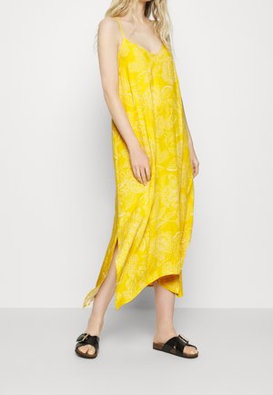 Kvinde iført en knaldgul blomstret slipkjole med tynde stropper og sorte sandaletter med spænde, poserer mod en ensfarvet hvid baggrund.