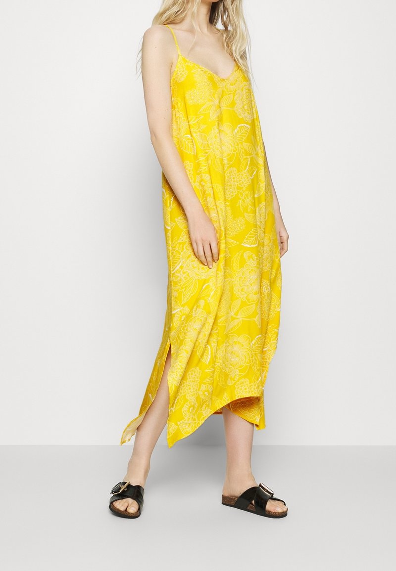 Femme portant une robe nuisette jaune vif à motifs floraux avec de fines bretelles et des sandales noires à boucle, posant devant un fond blanc uni.