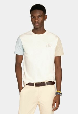 Giovane uomo che indossa una maglietta color crema con maniche azzurro chiaro e beige, pantaloni beige, cintura marrone e orologio da polso con cinturino giallo.