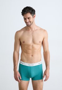 Teal bomuldsboxer med en hvid Calvin Klein logo-talje, tætsiddende design og glat tekstur. Modellen står med et neutralt udtryk.