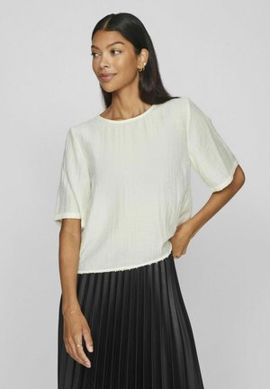 Blusa corta de manga corta en color crema con un acabado texturizado, escote redondo y dobladillo con ondas, combinada con una falda negra plisada.