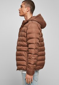 Bruine pufferjack met een capuchon, ribgebreide manchetten en horizontale quilt. Lichtgewicht materiaal met een gladde afwerking en een ritssluiting aan de voorkant.