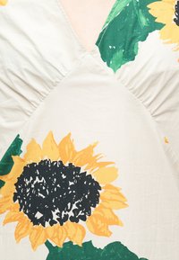 Ύφασμα σε κρεμ χρώμα με μεγάλο κίτρινο τύπωμα ηλίανθου και πράσινα φύλλα, με V-λαιμόκοψη και λεπτομέρειες με ραφή που συγκεντρώνεται.