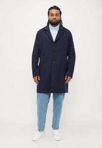 Trench in lana blu navy con tre bottoni, colletto a risvolto, tasche laterali, indossato sopra una polo azzurro chiaro e jeans azzurri, abbinato a sneakers bianche.