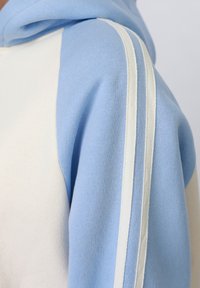 Sweat à capuche bleu clair et blanc avec deux bandes blanches courant le long du haut de la manche.