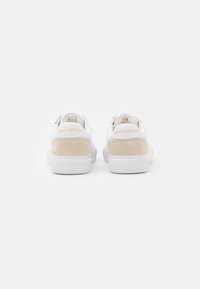 Ett par vita låga sneakers med beige mocka hälar och "Marc O’Polo"-logotyp i gul på baksidan.