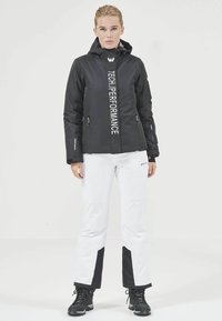 Schwarze isolierte Jacke mit "TECH PERFORMANCE" in vertikaler Schrift, kombiniert mit weißen Hosen mit schwarzen Akzenten und schwarzen Stiefeln.
