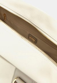 Interno di una borsa tote color crema con zip dorata, rivestimento beige e una piccola tasca con zip. Etichetta del marchio "GUESS" visibile all'interno.