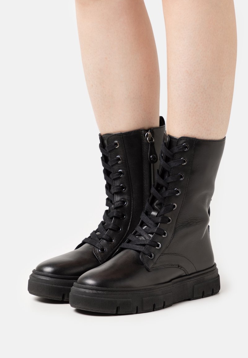 Bottes de combat noires en cuir à lacets avec fermeture éclair latérale portées sur des jambes nues, présentées sur un fond blanc uni.