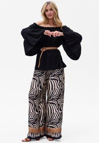 Schwarzes Off-Shoulder-Oberteil mit Glockenärmeln, kombiniert mit weiten Hosen im beigen und schwarzen Zebra-Muster. Braunem Gürtel als Akzent.