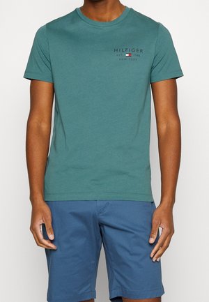 Mężczyzna w turkusowej koszulce Hilfiger z logo na klatce piersiowej i niebieskich szortach, stojący na tle jednolitego jasnego tła. Widoczne górna część ciała i nogi.