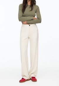 Beige wijde broek gemaakt van zachte stof met voorzakken en een enkele knoopsluiting, gecombineerd met rode sneakers en een aansluitend olijfgroen longsleeve shirt.