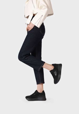 Persona che indossa jeans blu scuro, sneakers nere con zeppa e una giacca bianca corta, in piedi con una gamba leggermente sollevata.