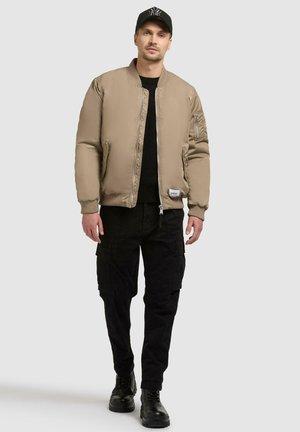 Giacca bomber beige con texture trapuntata, polsini a coste, tasche con zip, abbinata a pantaloni cargo neri e stivali neri. Design minimalista.