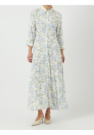 Femme portant une longue robe blanche à fleurs bleues, manches longues, et des chaussures à talons beiges sur un fond uni.