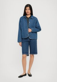 Tânără purtând o jachetă albastră matlasată, un tricou cu dungi, pantaloni scurți din denim și balerini negri, stând în fața unui fundal alb.