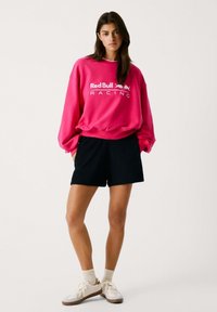 Sudadera de color rosa brillante con el logo blanco "Red Bull Racing", combinada con pantalones cortos negros y deportivas blancas. Ajuste casual y relajado. Textura suave.