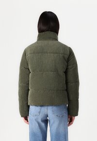 Olivově zelená kordurová péřová bunda s vysokým límcem, horizontálním prošíváním a hladkou texturou. Spárovaná se světle modrými džíny.