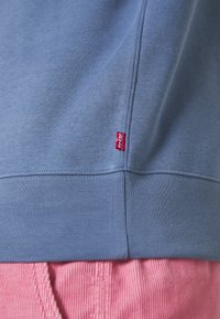 Sweatshirt blu con orlo e piccolo tag rosso Levi's sopra pantaloni in corduroy rosa, che mostrano la texture del tessuto e i dettagli delle cuciture.