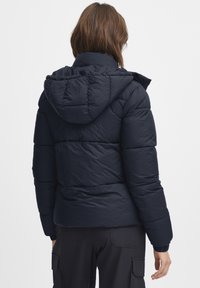 Marineblauwe pufferjack met capuchon, voorzien van een doorgestikte textuur en elastische manchetten. Ontworpen voor warmte met een aansluitende pasvorm. Zwart voering zichtbaar.