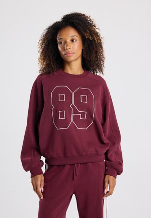 Bordeaux sweatshirt met witte omranding van het getal "89" aan de voorkant, zacht materiaal, relaxte pasvorm, geribde boorden en zoom. Draagt met bijpassende broek.