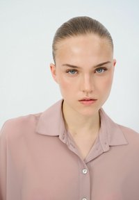 Blusa trasparente di un delicato rosa chiaro con colletto classico e sottili dettagli sui bottoni, caratterizzata da una texture morbida e un design minimalista.