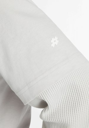 Primo piano di una camicia bianca a maniche lunghe a strati con una manica interna testurizzata a maglia a nido d'ape e un piccolo simbolo hashtag sulla manica esterna.