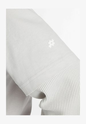 Primo piano di una camicia bianca a maniche lunghe a strati con una manica interna testurizzata a maglia a nido d'ape e un piccolo simbolo hashtag sulla manica esterna.