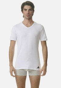 Vit bomulls-T-shirt med V-ringning och korta ärmar, med en subtil Adidas-logotyp på vänster sida. Modell iförd gråa kalsonger.