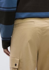 Beežid tööpüksid küljetasku ja tagumise läbilöögitaskuga, millele on lisatud mitmevärviline kootud kampsun siniste ja mustade triipudega.