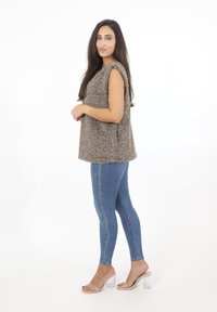 Grå fuskpäls utan ärmar, mjuk textur; kombinerad med åtsittande blå denimjeans och klara blockklacksandaler. Enkel, modern silhuett.