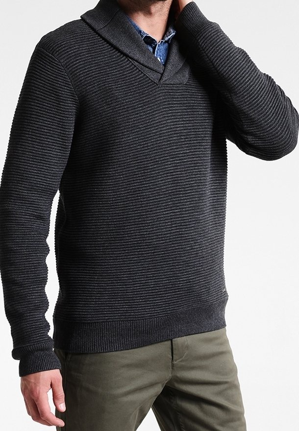 Mężczyzna w ciemnoszarym swetrze z prążkowanym splotem, założonym na niebieską koszulę z kołnierzykiem, w oliwkowych spodniach, z ręką uniesioną do szyi.