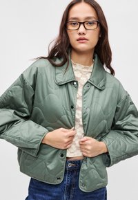 Μίντ πρασινή επενδυμένη ζακέτα με κλείσιμο από κουμπιά και μεγάλο γιακά. Φορεμένη πάνω από μια κρέμα δαντελένια μπλούζα, με χαλαρή εφαρμογή και μαλακή υφή.