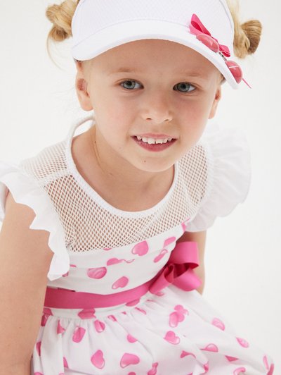 Vestido blanco con patrones de corazones rosas, escote de malla y mangas con volantes; complementado con una visera blanca con un lazo rosa.