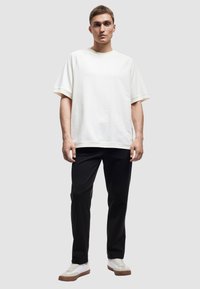 Lässig geschnittenes T-Shirt aus cremefarbener Baumwolle mit übergroßen Ärmeln, kombiniert mit schwarzen Hosen und weißen Sneakers mit hellbraunen Sohlen.