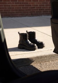 Paire de bottes d'hiver noires avec semelles épaisses sur un trottoir ensoleillé près d'un mur en briques et d'une portière de voiture ouverte.
